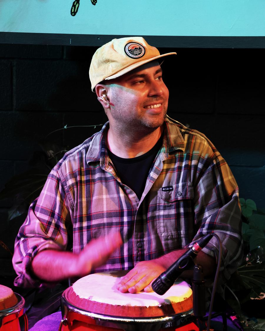 Eric Mendez in a plaid shirt and cap, smiling as he plays the congas under pink and blue stage light.